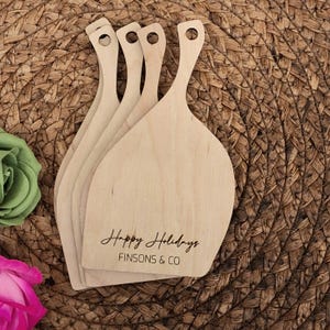 May include: A stack of five light-colored wooden serving boards with handles. Each board has a rounded paddle shape and a small hole in the handle. The words "Happy Holidays" and "FINSONS & CO" are printed on the front.