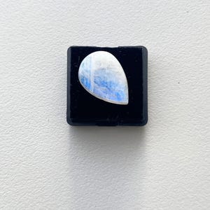 Può includere: Una pietra di luna lucidata a forma di pera, con una sfumatura di colore bianco e blu. La pietra è posta su un piccolo espositore quadrato nero. La gemma viene probabilmente utilizzata per gioielli o come oggetto decorativo.