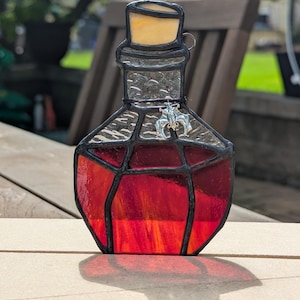 May include: Stained glass bottle with a red and orange liquid. The bottle has a black outline and a cork stopper. A silver charm is attached to the front. The bottle is sitting on a wooden surface.