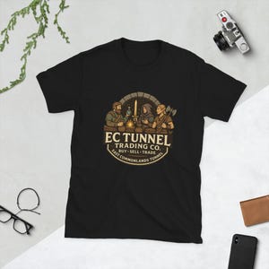 May include: Black t-shirt featuring a circular graphic with three figures around a table, a sword, and the text "EC TUNNEL TRADING CO." in a vintage style. The design includes the words "BUY SELL TRADE" and "EAST COMMONLANDS TUNNEL".