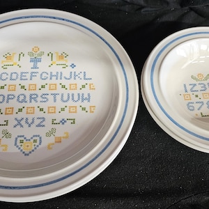 May include: A white ceramic dinner plate and a matching bowl, both featuring blue and yellow cross-stitch style patterns. The plate displays the alphabet from A to Z, while the bowl features the numbers 1 through 0, each accented with small floral and geometric motifs and blue rim bands.