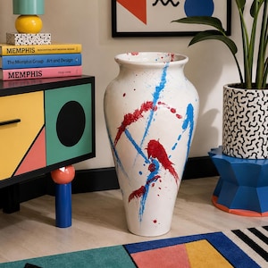 May include: A tall floor vase with a white base featuring a decorative splatter pattern in bright red and blue paint. The vase has a wide, rounded rim that tapers down to a narrow base. The text 16" TALL FLOOR VASE is printed at the bottom.