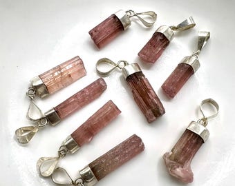 May include: A collection of pink tourmaline pendants with silver-colored metal caps and loops. The raw, cylindrical gemstones vary in shades of pink and brown. The pendants are displayed on a white surface.