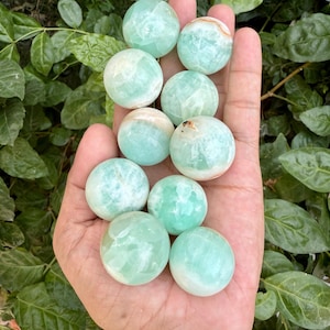 May include: A hand displays a collection of polished, spherical gemstones. The stones are a soft, translucent green with colour and texture variations. The spheres are set against green foliage, creating a natural and earthy aesthetic.