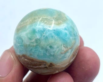May include: A polished sphere of larimar gemstone, held in a hand. The stone features a blend of light blue, turquoise, and brown hues, with a swirling pattern. The sphere is smooth and round, showcasing the natural beauty of the mineral.