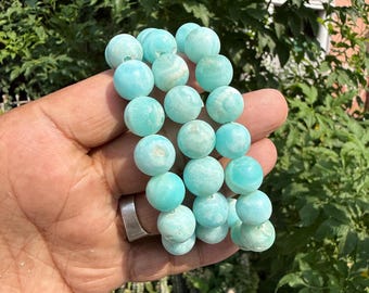 Könnte beinhalten: Ein Armband mit mehreren runden, hellblauen Perlen. Die Perlen haben ein marmoriertes Aussehen und sind aufgefädelt. Das Armband wird in einer Hand gehalten, wobei ein silberner Ring sichtbar ist. Der Hintergrund ist verschwommen mit grünem Laub.