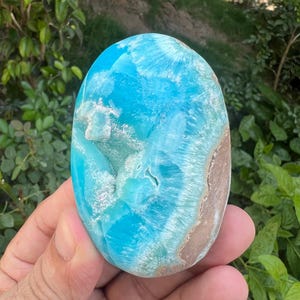 May include: A polished, oval gemstone with a striking blue and white pattern, reminiscent of ocean waves. The stone has a smooth, glossy surface and a band of brown along one edge. Held in a hand against a backdrop of green foliage.