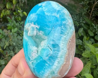 May include: A polished, oval-shaped gemstone with a striking blue and white pattern, reminiscent of ocean waves. The stone has a smooth, glossy surface and a band of brown along one edge. Held in a hand against a backdrop of green foliage.