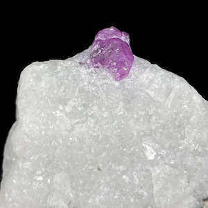 Pode incluir: Um close-up de um espécime mineral bruto. O corpo principal é uma grande rocha branca e cristalina. Um pequeno cristal rosa vibrante está no topo. O fundo é preto sólido.