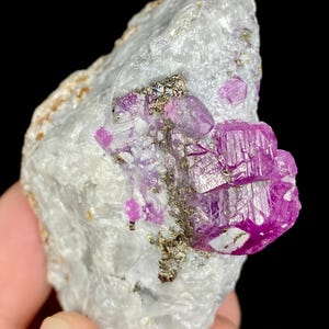May include: A close-up of a rough, white rock formation with vibrant, hot pink crystal clusters. The crystals have a prismatic structure and are contrasted by small, metallic flecks.
