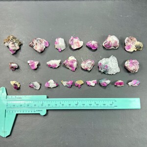 Puede incluir: Una colección de piedras preciosas de rubí en bruto de varios tamaños y formas, que muestran un vibrante color rosa y rojo. Las piedras están sobre un fondo oscuro, con una regla verde para la escala. Las piedras preciosas son opacas y translúcidas.