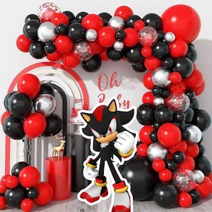 Op de afbeelding: Een feestelijke ballonnenboog in rood, zwart en zilver omringt een stripfiguur. De woorden "Oh Baby" zijn zichtbaar op de muur. Een taart en een gouden hertenbeeldje zijn ook op de achtergrond te zien. De ballonnen zijn in een boog gerangschikt.