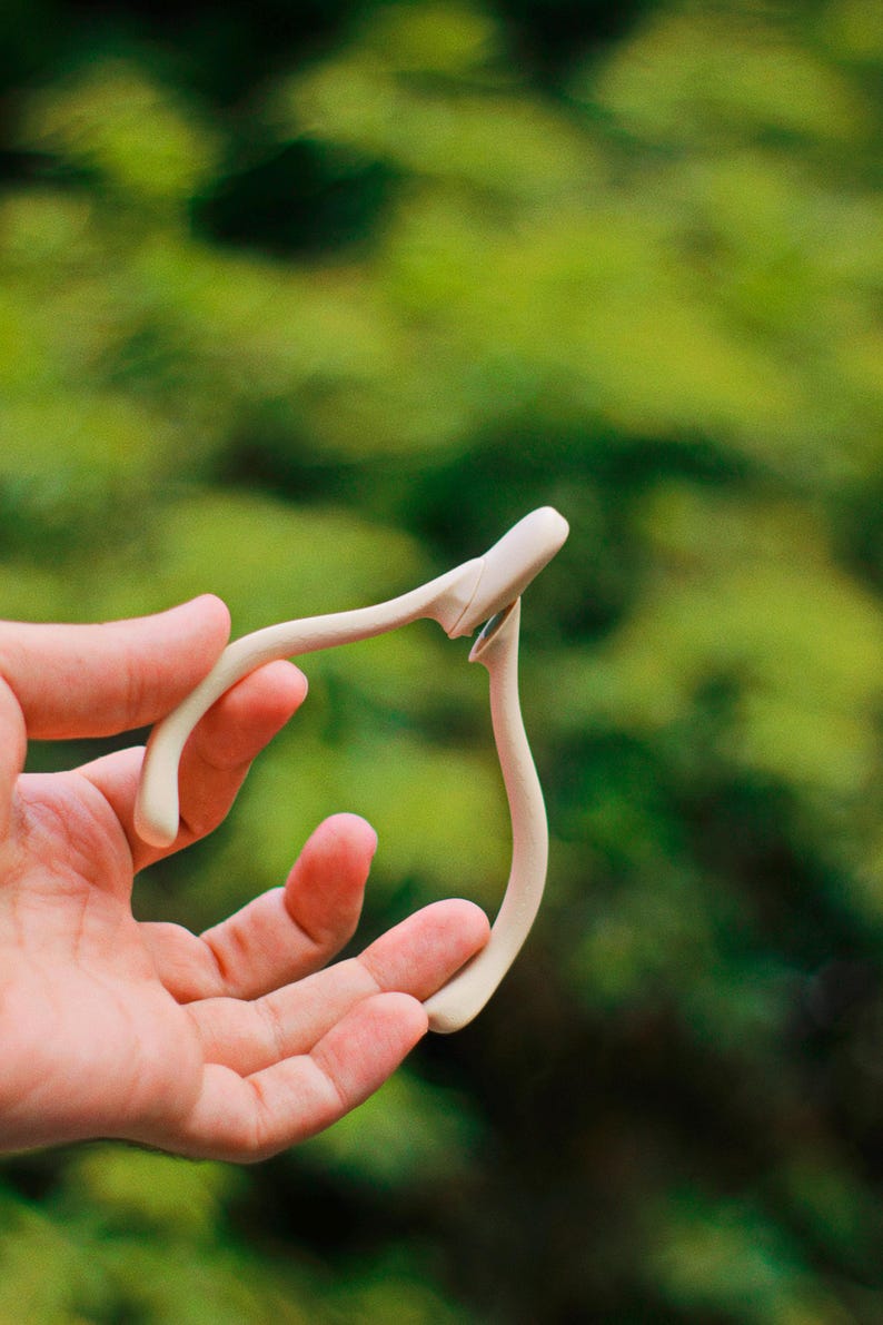 May include: A cream-colored, wishbone-shaped object is held in a hand against a blurred green background. The object has a curved design with a small, flat top and two elongated, curved arms.