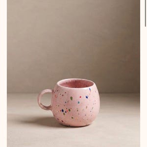 Peut inclure: Une tasse en céramique ronde d'un ton rose pastel doux, ornée d'un motif moucheté composé de petits points multicolores dans des nuances de bleu, de vert et d'orange sur toute sa surface extérieure. Elle possède une anse incurvée et arrondie.