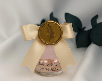 May include: A small glass jar filled with pink beads, topped with a cream-colored ribbon and a gold-colored wax seal. The seal features a leaf design. The jar has the names "Malice & Oliv" and the date "28.08.2025" printed on it.