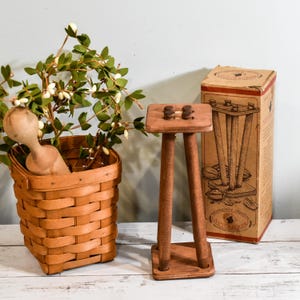 Może przedstawiać: Drewniany stojak na dziadka do orzechów z kwadratową górą i podstawą, wsparty na dwóch cylindrycznych nogach. Za stojakiem znajduje się zabytkowe pudełko z ilustracją dziadka do orzechów. Po lewej stronie znajduje się pleciony kosz z drewnianym tłuczkiem i zielenią.