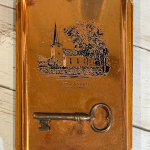 May include: A rectangular copper-colored plaque with a church illustration and the text "SAUDA KYRKJE anno 1866." A vintage key is attached to the front. The plaque is framed with wood.