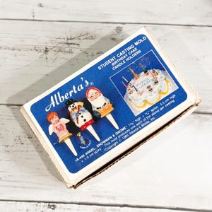 Puede incluir: Una caja vintage de Alberta's Student Casting Mold para portavelas de pastel de cumpleaños. La caja presenta un diseño azul y blanco con ilustraciones de ángel, muñeco de nieve y gnomo. Los adornos miden aproximadamente 3,8 cm de alto.