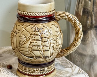 May include: A beige ceramic mug with a nautical theme. The mug features a detailed relief of a sailing ship and buildings. The handle is rope-like, and the base has text. The mug has a cream-colored interior and is approximately 5 inches tall.