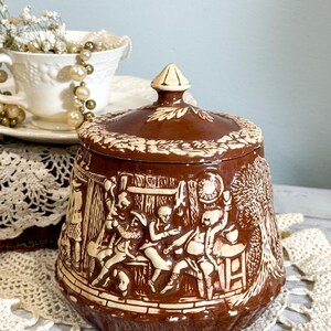 May include: Brown ceramic jar with a lid, featuring a raised relief design of a gathering scene. The jar has a cream-colored relief design against a brown background. The lid has a decorative finial.