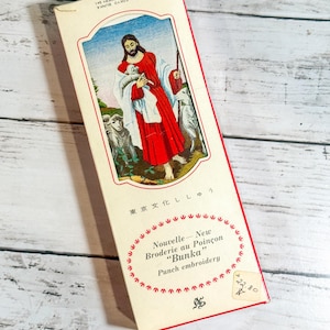 Könnte beinhalten: Vintage Stickpackungskarton mit religiöser Illustration. Die Box hat ein rot-weißes Farbschema mit Text in Englisch und Japanisch. Das Bild auf der Box zeigt eine Figur in rotem Gewand mit Schafen.