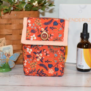 May include: A small, square fabric box with a floral pattern in orange, white, and blue. The box has a peach-colored trim and a wooden button closure. A brown glass bottle with a dropper is to the right.