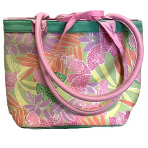 May include: A colorful tote bag with a frog and leaf pattern. The bag features a pink bow, handles, and trim. The background is a mix of green, yellow, and orange, with pink frogs and purple leaves. The handles are made of pink leather.