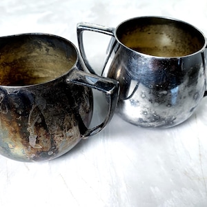 May include: Two antique silver-plated cream and sugar bowls. The bowls have a rounded shape with a handle on each side. The silver plating shows signs of wear and discoloration, giving them a vintage appearance. The interior of the bowls is a tarnished gold color.
