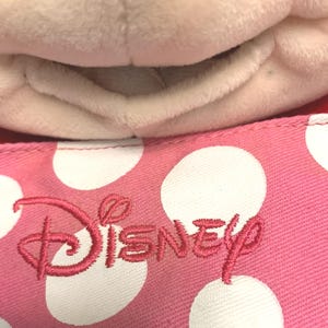 May include: A plush toy with a soft white fabric texture positioned above a pink fabric surface featuring a white polka dot pattern and the word Disney embroidered in pink thread. The textile has a visible woven grain and a stitched seam edge.