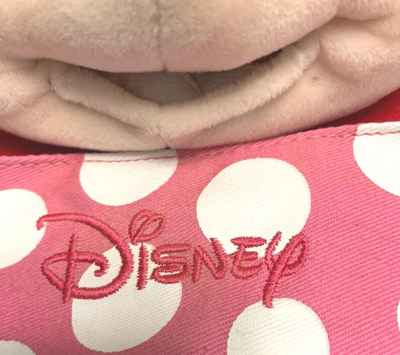 May include: A plush toy with a soft white fabric texture positioned above a pink fabric surface featuring a white polka dot pattern and the word Disney embroidered in pink thread. The textile has a visible woven grain and a stitched seam edge.