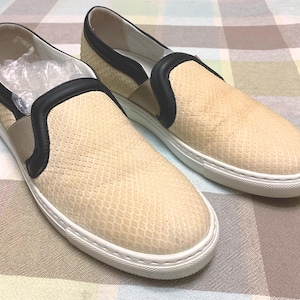 May include: A pair of slip-on sneakers featuring a beige snakeskin-textured upper, black leather trim around the collar and elastic side panels, and a thick white rubber sole. The interior of the left shoe contains a clear plastic insert.