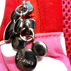 May include: A bright red zipper closure on a fabric item featuring a metallic silver charm shaped like a mouse head. The charm has three circular parts and the word Disney embossed on the bottom. The surrounding fabric is pink with white dots.