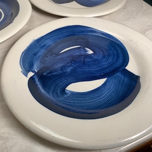 May include: White ceramic plates with a bold, abstract design in shades of blue. The plates feature a hand-painted, swirled pattern, creating a modern and artistic aesthetic. The plates are suitable for serving food.