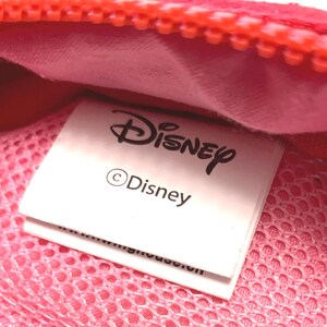 May include: A bright red zippered pouch featuring a pink mesh interior lining. A white fabric tag is sewn inside, clearly displaying the Disney logo in black text, followed by the copyright symbol and the word Disney in a smaller font size.