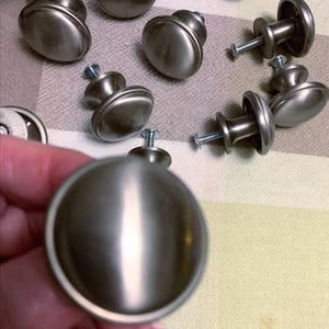May include: A collection of round cabinet knobs with a brushed silver finish. Each knob features a smooth, domed surface and a central stem with an attached metal screw for installation. The knobs have a metallic sheen and a circular profile.