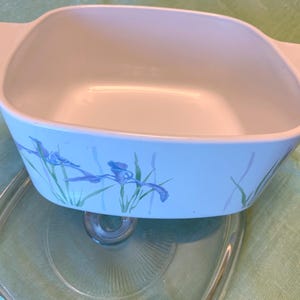 May include: A square, white casserole dish with a light blue floral design featuring blue irises and green stems. The dish has two handles and sits atop a clear glass lid. A vintage kitchenware item.