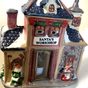 May include: A ceramic Christmas night light shaped like Santa's Workshop. The building has a red and brown facade, a dark blue roof with snow, and the text "SANTA'S WORKSHOP" above the window. The night light has a chimney and a small figure of Santa.