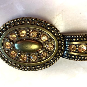 May include: An oval-shaped, antique gold-tone belt buckle with a decorative design. The buckle features a central oval with amber-colored rhinestones and a rope-like border. The attached belt loop also has amber rhinestones.