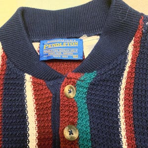 May include: A knit polo shirt featuring vertical stripes in navy blue, burgundy, white, and teal. The shirt has a navy blue ribbed collar, a button-up placket with two metallic buttons, and a blue Pendleton label at the neck marked with size XL.