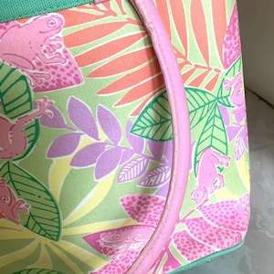 May include: A pink and green tote bag with a tropical frog print. The bag features a pink handle and a green trim. The pattern includes pink frogs, green leaves, and orange and purple foliage. The bag is made of a durable fabric.
