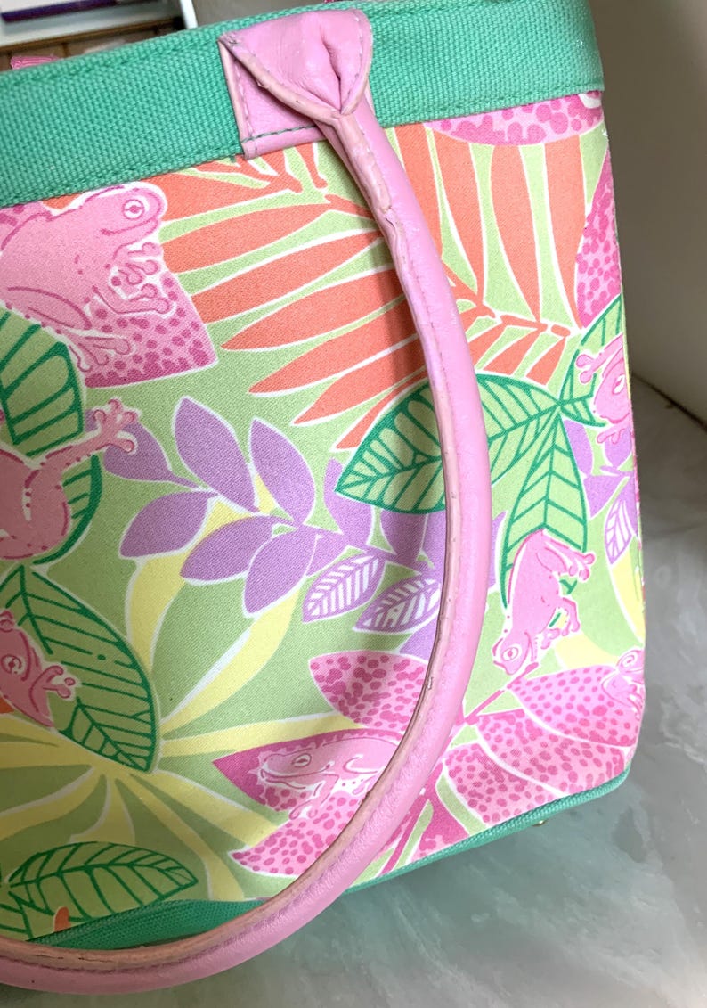 May include: A pink and green tote bag with a tropical frog print. The bag features a pink handle and a green trim. The pattern includes pink frogs, green leaves, and orange and purple foliage. The bag is made of a durable fabric.