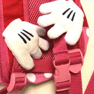 May include: A pink and red backpack featuring two plush white cartoon-style gloves attached to the shoulder straps. The backpack includes pink adjustable plastic buckles, a mesh back panel, and a fabric section with white polka dots on a pink base.