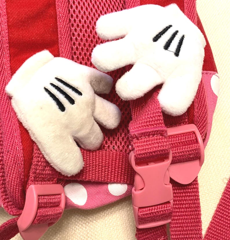 May include: A pink and red backpack featuring two plush white cartoon-style gloves attached to the shoulder straps. The backpack includes pink adjustable plastic buckles, a mesh back panel, and a fabric section with white polka dots on a pink base.