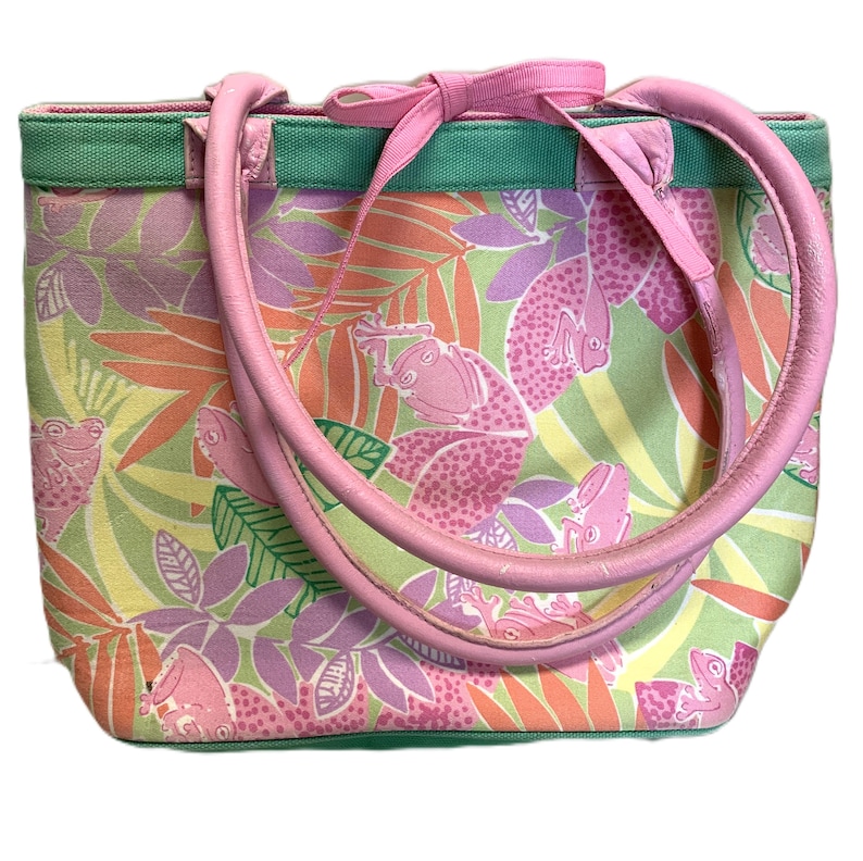 May include: A colorful tote bag with a frog and leaf pattern. The bag features a pink bow, handles, and trim. The background is a mix of green, yellow, and orange, with pink frogs and purple leaves. The handles are made of pink leather.