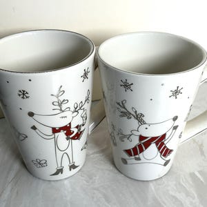 May include: Two white ceramic mugs with a silver rim and a handle. Each mug features a cartoon reindeer wearing a red and white scarf, with snowflakes and other festive designs. The mugs are ideal for hot beverages.