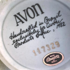 May include: The underside of a ceramic object featuring the black printed text AVON, Handcrafted in Brazil exclusively for Avon Products Inc., 1982. Below this is the gold stamped number 117329 and an oval black sticker reading Made Etamarte in Brazil.