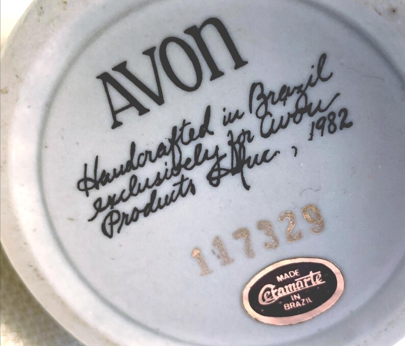 May include: The underside of a ceramic object featuring the black printed text AVON, Handcrafted in Brazil exclusively for Avon Products Inc., 1982. Below this is the gold stamped number 117329 and an oval black sticker reading Made Etamarte in Brazil.