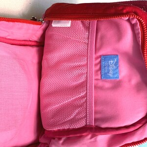 May include: A red backpack with a bright pink interior lining. The inside features a pink mesh pocket and a small blue rectangular fabric tag with the word Disney printed in white text. The bag has a red zipper closure along the top edge.