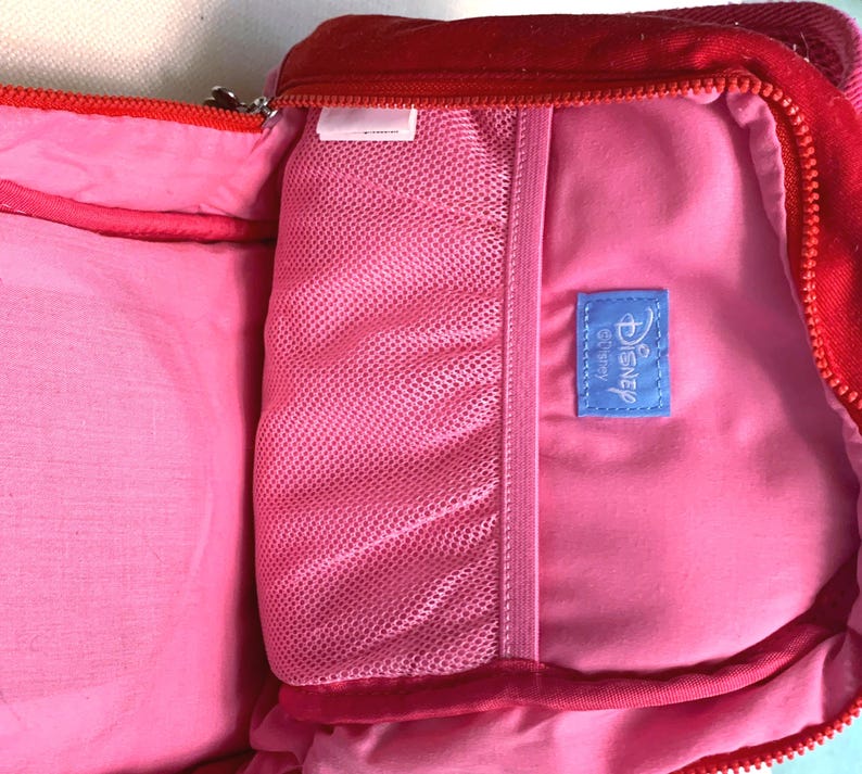 May include: A red backpack with a bright pink interior lining. The inside features a pink mesh pocket and a small blue rectangular fabric tag with the word Disney printed in white text. The bag has a red zipper closure along the top edge.