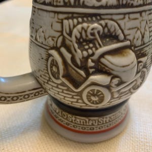 May include: A ceramic beer stein featuring a raised relief design of a vintage automobile on a brick-patterned background. The base of the stein includes the text 1910 Stanley Steamer, with a handle decorated in a geometric pattern and brown accents.